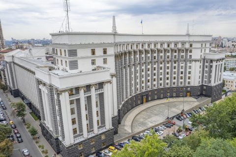 Велика біла урядова будівля з колонами та прапорами України в Києві