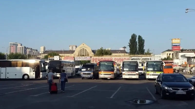 Пасажири з валізою йдуть між рядами автобусів на автовокзалі